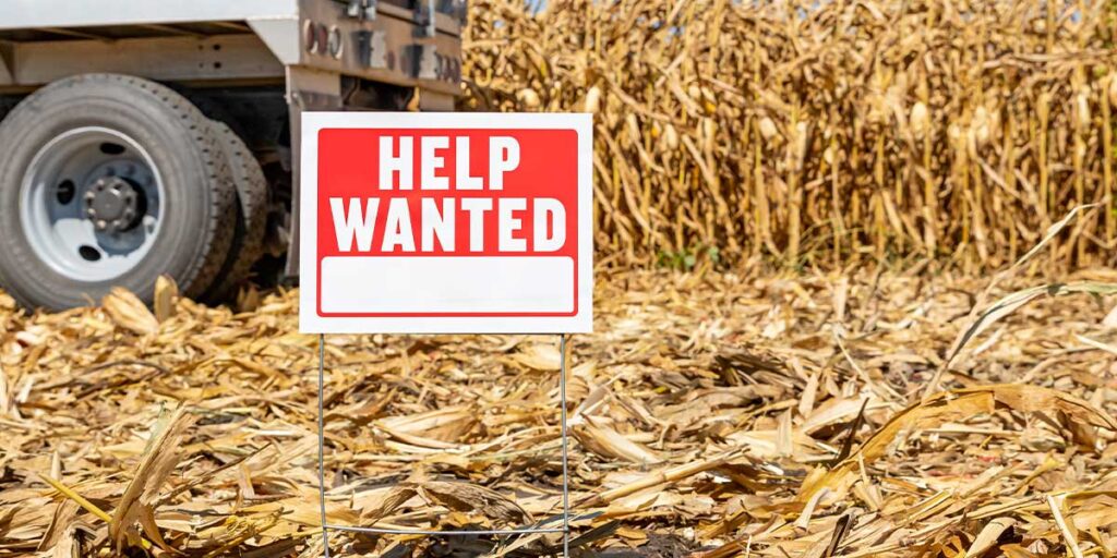 This is a picture of a help wanted sign in front of a tractor and bales of hay.